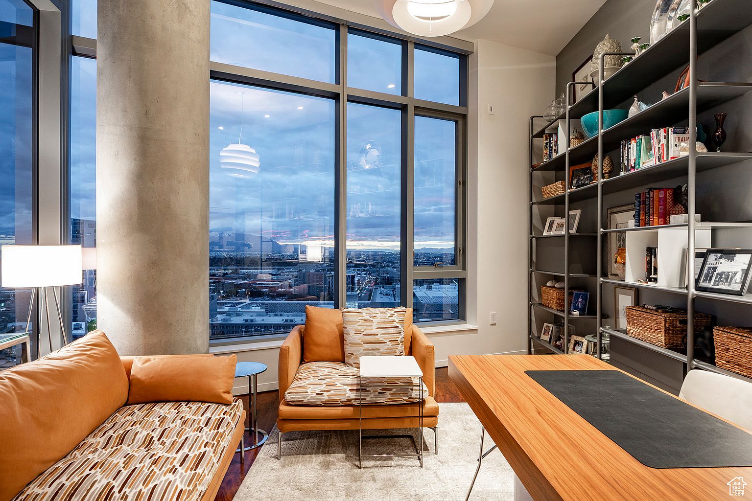 This is an interior shot of a modern office space featuring a large window with a city view. The room includes a comfortable seating area with an orange couch and chair, a wooden desk with a black desk pad, and a tall bookshelf filled with books and decorative items. The overall impression is stylish and sophisticated, perfect for a professional working from home.