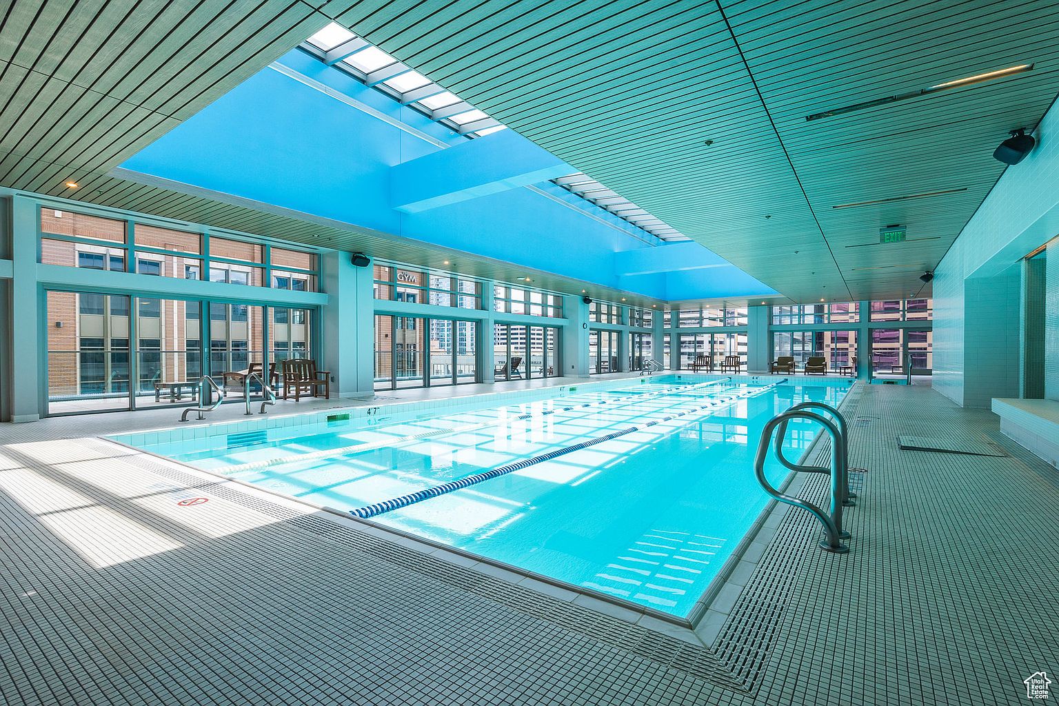 This image showcases a well-lit indoor swimming pool area, featuring a large pool with lane markers, surrounded by tiled flooring and floor-to-ceiling windows that offer city views. The space is illuminated by natural light from a skylight, creating a bright and inviting atmosphere, perfect for relaxation and exercise.