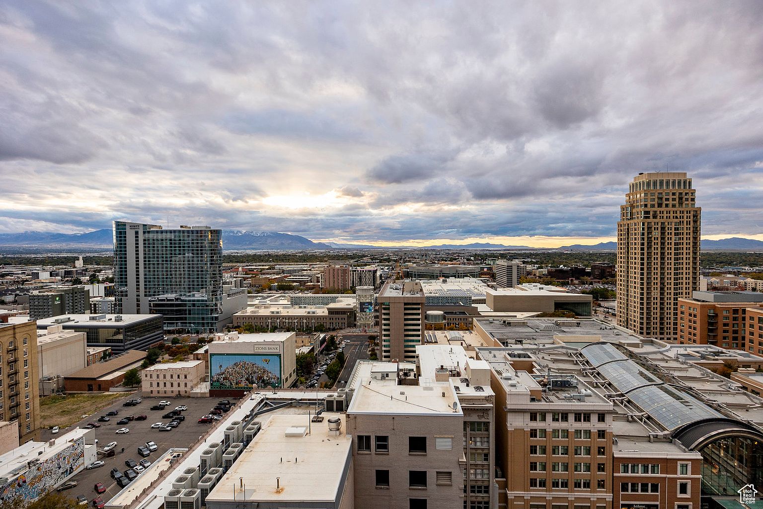 This aerial view showcases a cityscape with a mix of modern high-rise buildings and older structures, under a dramatic cloudy sky. The composition highlights the urban density and architectural diversity, with distant mountains providing a scenic backdrop. The image conveys a sense of scale and urban sophistication, suitable for showcasing the location and surroundings of a property.