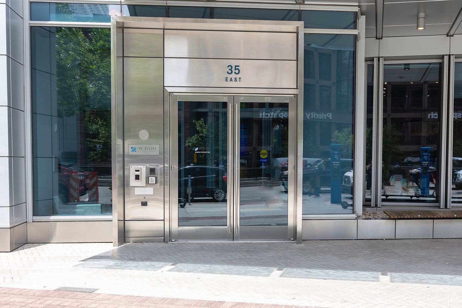 The image showcases the modern entryway of a building, featuring a stainless steel facade with the address "35 East" prominently displayed above the double glass doors. A security panel is visible on the left, and the reflective glass surfaces create a sleek and professional aesthetic. The entrance is framed by large windows, offering glimpses of the surrounding urban environment.