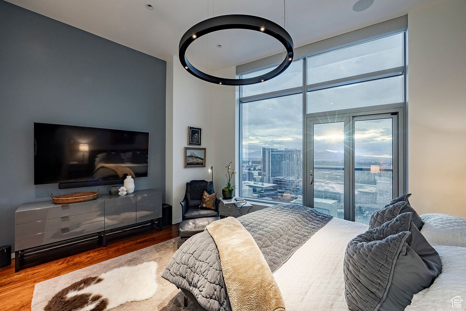 This is a luxurious primary bedroom featuring a large window with a city view, a modern circular chandelier, and a gray accent wall with a mounted TV. The room is furnished with a gray dresser, a leather chair, and a plush bed with gray and white bedding. The overall impression is one of sophistication and comfort.