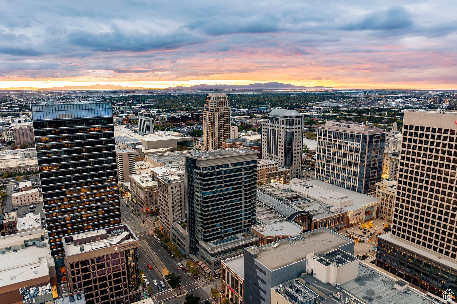 This aerial view showcases a vibrant cityscape at sunset, featuring a mix of modern skyscrapers and older buildings. The warm hues of the setting sun paint the sky, creating a dramatic backdrop for the urban landscape. Distant mountains add depth to the scene, emphasizing the city's location and surroundings.