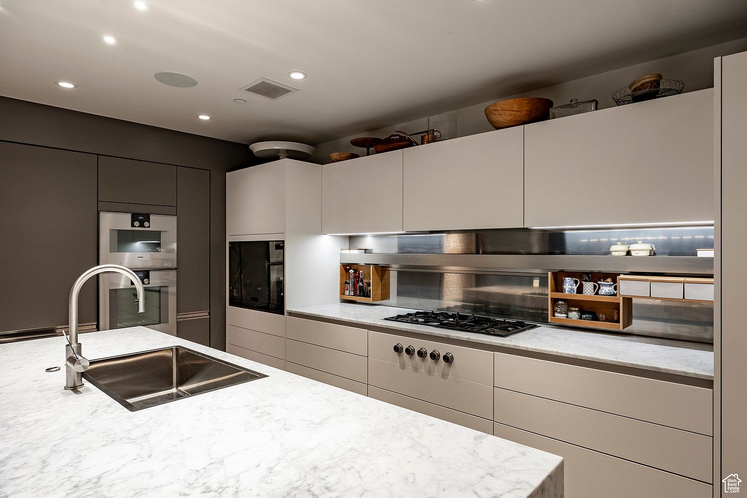 This is a modern kitchen featuring a large island with a marble countertop and a stainless steel sink. The kitchen is equipped with built-in stainless steel appliances and sleek cabinetry in neutral tones. The design emphasizes clean lines and a minimalist aesthetic, creating a sophisticated and functional space.