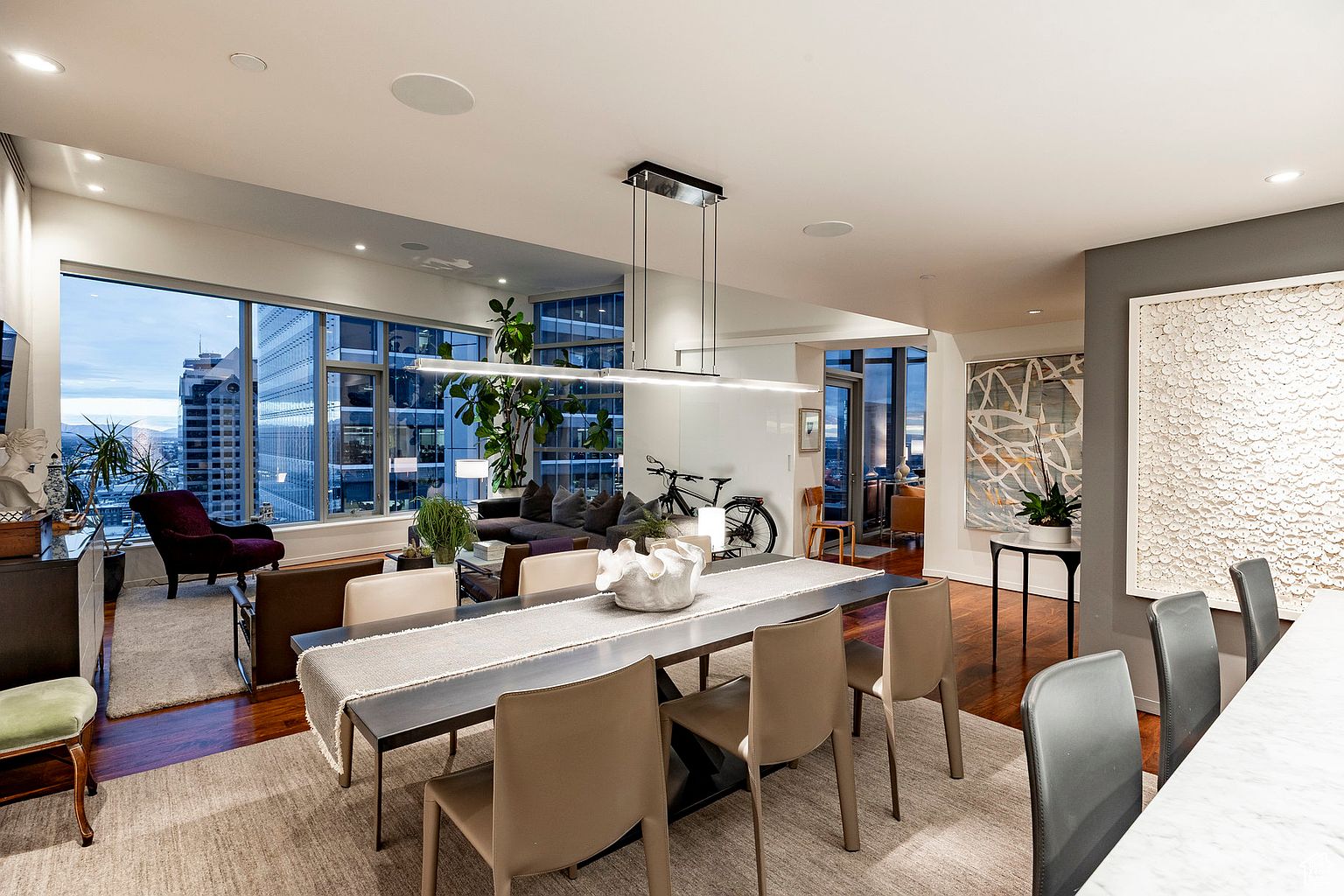 This interior shot showcases a modern dining room with a long, dark wood table set for dining, complemented by tan chairs and a light-colored runner. Large windows offer a stunning city view, while contemporary art and a unique light fixture add to the room's sophisticated ambiance. The space is open and airy, blending seamlessly with the living area.