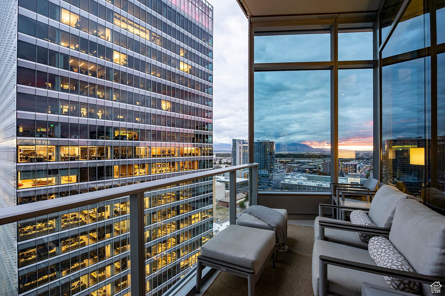 This image showcases a luxurious balcony with modern outdoor furniture, including cushioned chairs and ottomans, set against a stunning cityscape backdrop. The view includes a neighboring high-rise building with illuminated office spaces and a distant mountain range at sunset, creating a sense of urban sophistication and expansive living.