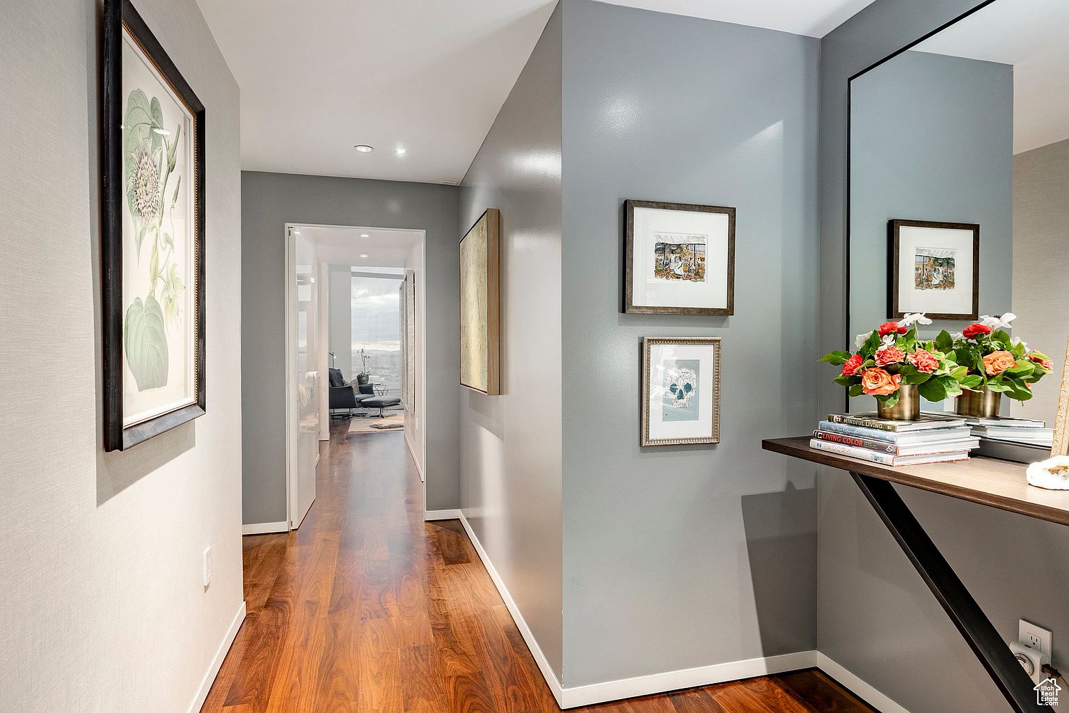 This is an interior shot of a hallway featuring hardwood floors and gray walls. Artwork adorns the walls, and a console table with floral arrangements and books adds a touch of elegance. The hallway leads to a bright living space, creating an inviting and stylish transition between rooms.
