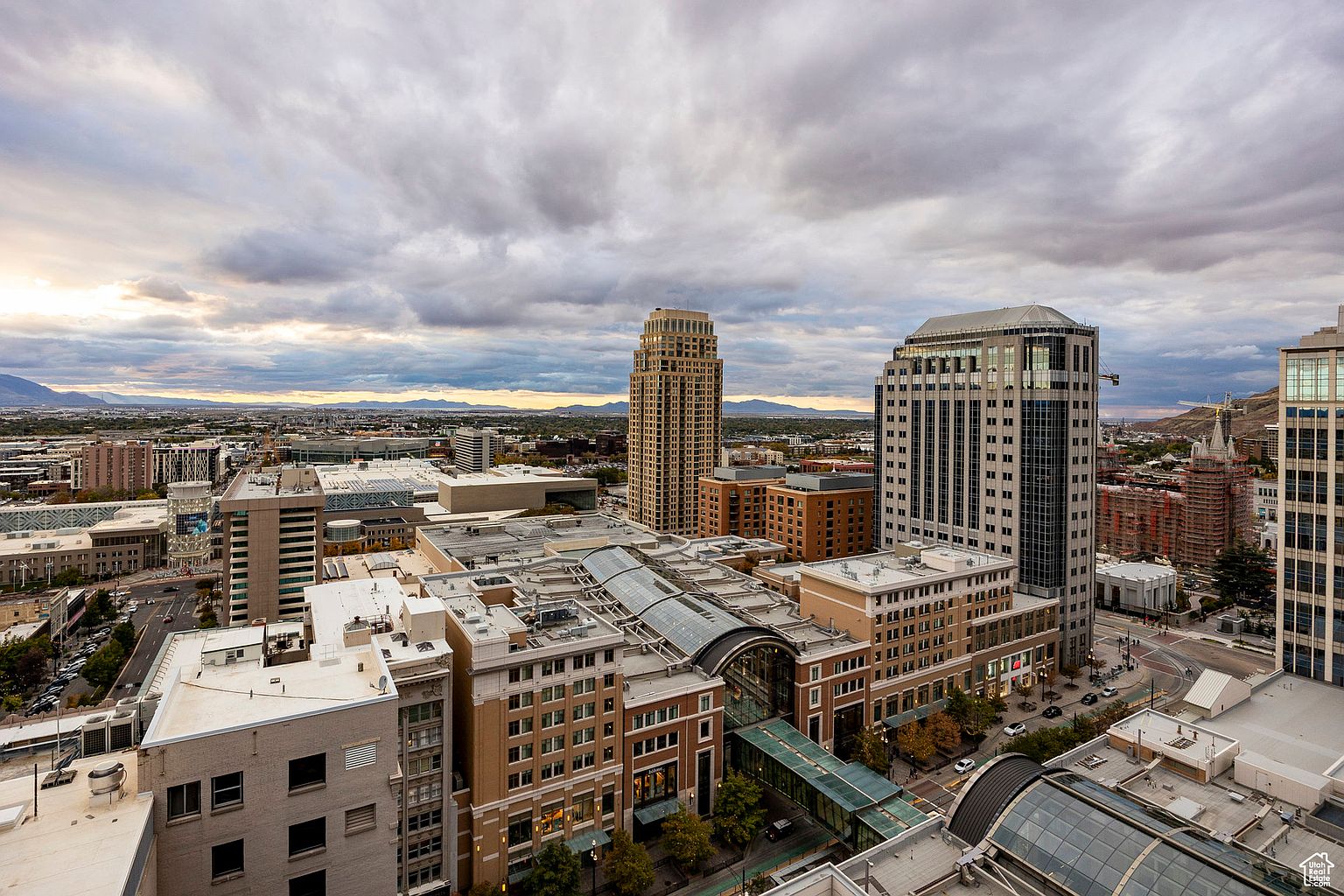 This aerial view showcases a vibrant cityscape with a mix of modern skyscrapers and commercial buildings under a cloudy sky. The architecture features a blend of glass and brick, with pedestrian walkways connecting various structures. The overall impression is one of a bustling urban center with a well-planned infrastructure.