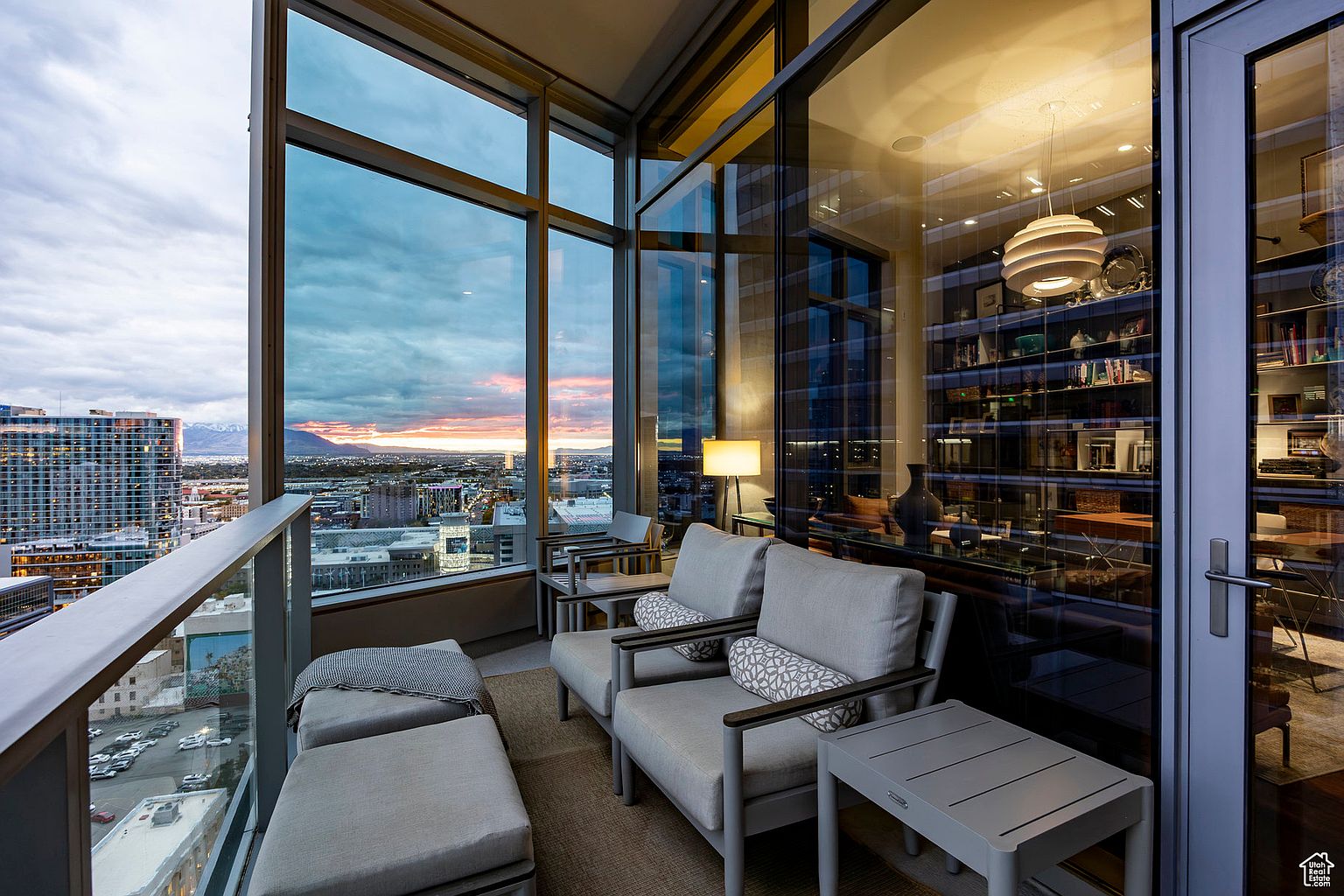 This image showcases a luxurious balcony with comfortable seating, offering stunning city views at sunset. The balcony features modern furniture, including cushioned chairs and ottomans, complemented by a sleek glass railing. The expansive windows provide a seamless transition between indoor and outdoor living, enhancing the appeal of this high-rise residence.