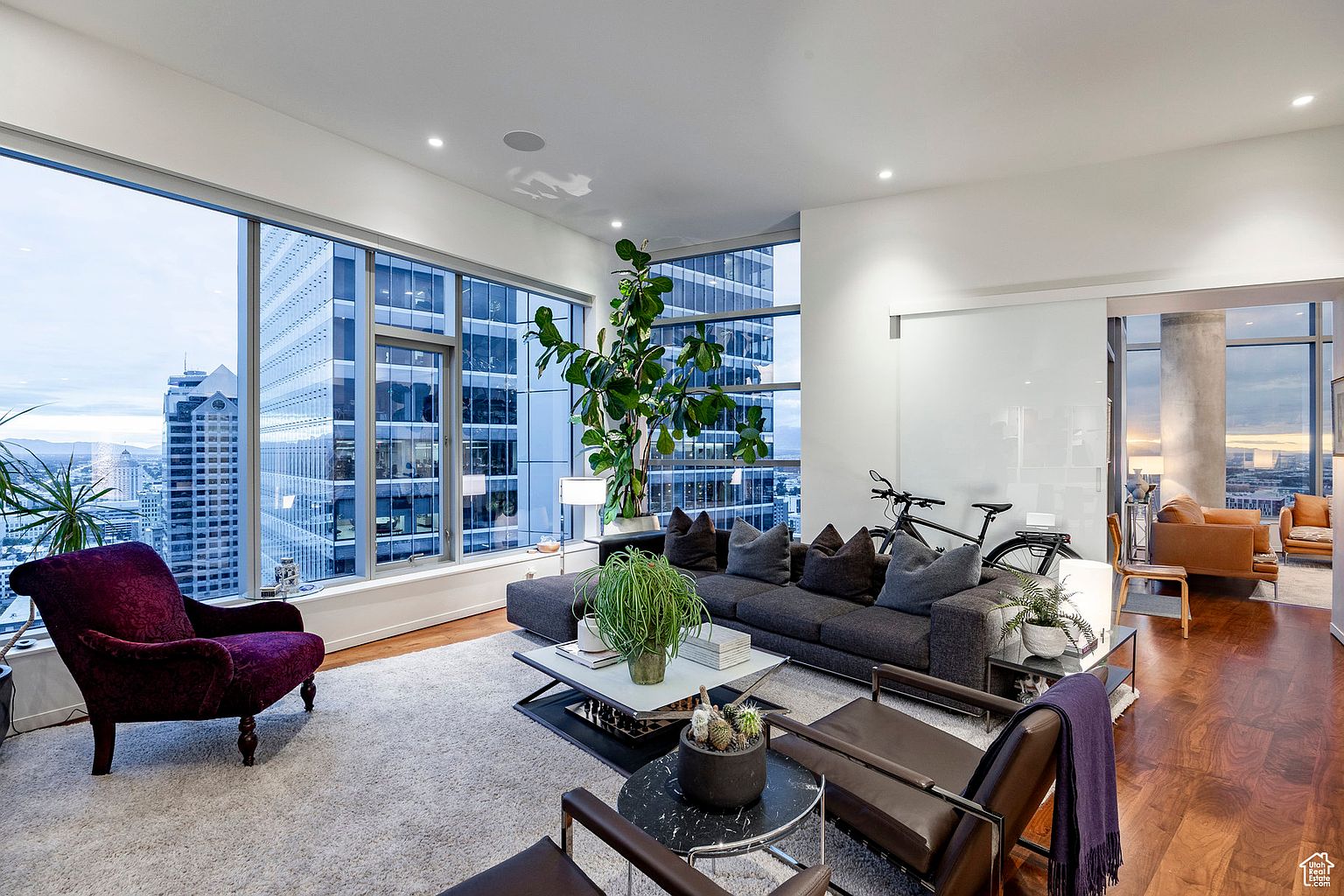 This is a luxurious living room in a high-rise apartment, featuring floor-to-ceiling windows with city views. The room is furnished with a large gray sectional sofa, a purple armchair, and modern coffee tables. Hardwood floors and a neutral-toned rug add warmth and texture to the space, while a bicycle is casually placed near the sliding glass door.