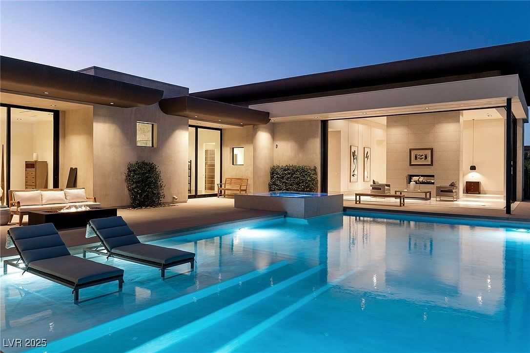 This image showcases a luxurious backyard pool and spa area at dusk. The pool features illuminated steps and clear blue water, reflecting the surrounding modern architecture. Lounge chairs are positioned near the pool, and the open-concept design seamlessly connects the indoor and outdoor living spaces, creating an inviting and relaxing atmosphere.