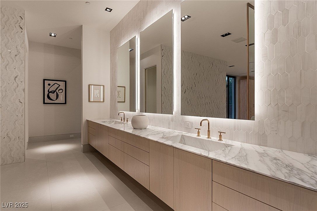 This is a luxurious primary bathroom featuring a long, light wood vanity with a marble countertop and dual sinks. Large, illuminated mirrors hang above the vanity, reflecting the modern design and enhancing the bright atmosphere. The walls are adorned with stylish tile, contributing to the bathroom's elegant and contemporary aesthetic.