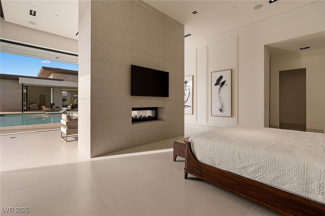 This is a luxurious primary bedroom featuring a modern design with a neutral color palette. A striking architectural element is the central wall with a built-in fireplace and mounted television, separating the sleeping area from an outdoor pool area visible through large sliding glass doors. The room is furnished with a sleek, low-profile bed and minimalist artwork, creating a serene and sophisticated atmosphere.