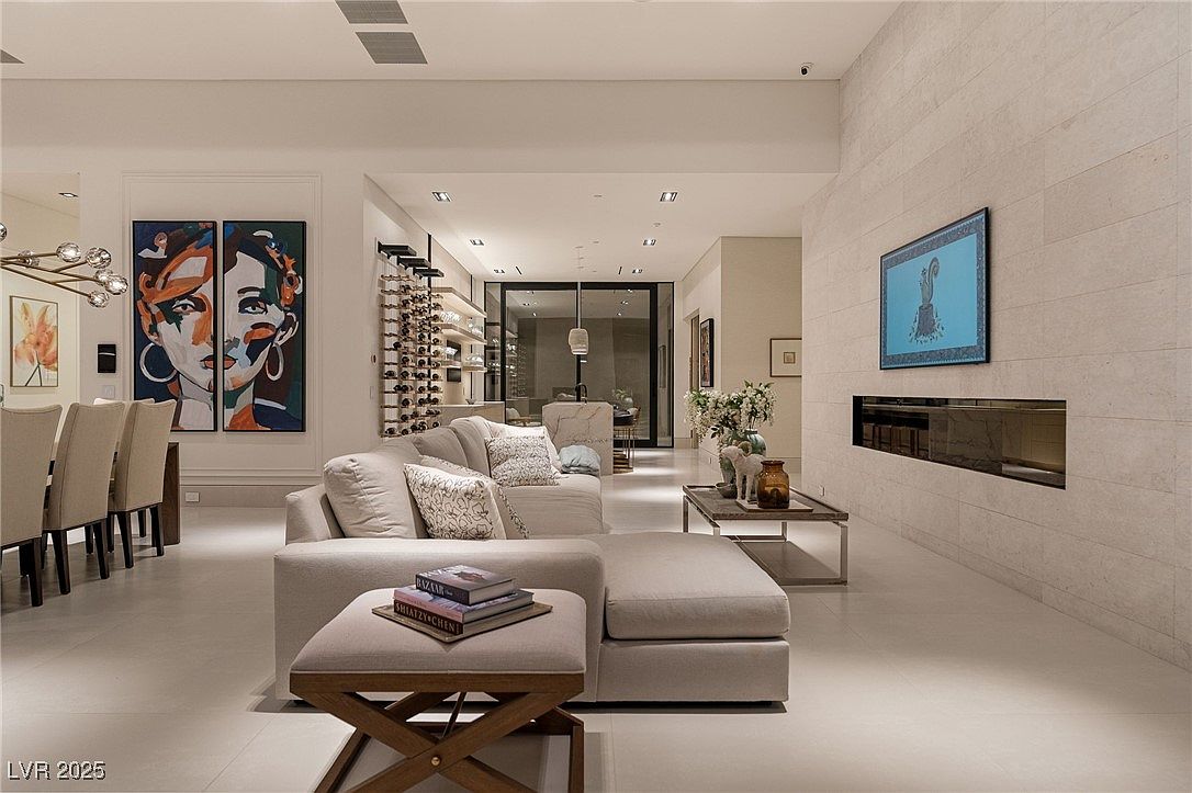 This interior shot showcases a modern living room with a neutral color palette, featuring a sectional sofa with decorative pillows, a contemporary coffee table, and a sleek fireplace with a mounted television. Artwork adorns the walls, and the open floor plan seamlessly connects the living area to the dining space, creating an inviting and sophisticated atmosphere. The perspective is from a low angle, emphasizing the spaciousness of the room.