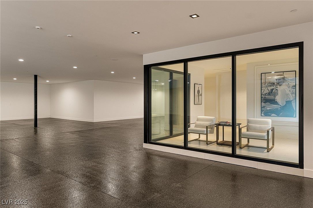 The image showcases a modern living room with a minimalist aesthetic. The room features a polished concrete floor, white walls, and a large glass partition that separates the space. Inside the glass partition, there are two armchairs and a small table, creating a cozy seating area with artwork on the wall.