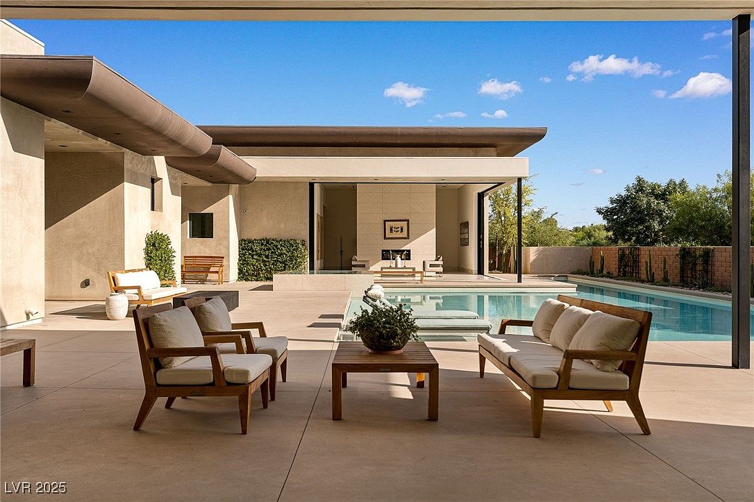 This image showcases a luxurious outdoor living space with a stunning pool and spa area. The patio is furnished with modern outdoor seating, including chairs and a sofa, arranged around a coffee table. The architecture features clean lines and a seamless transition between indoor and outdoor spaces, creating an inviting and relaxing atmosphere.