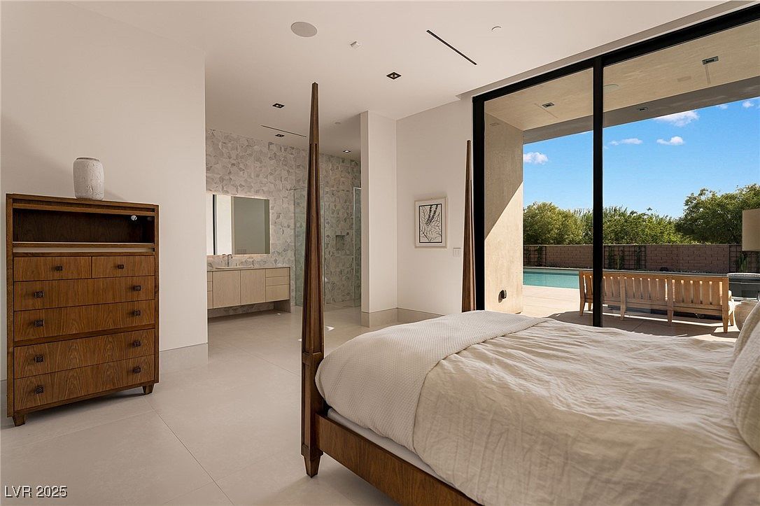 This is a primary bedroom featuring a four-poster bed with neutral bedding, a wooden dresser, and an en-suite bathroom visible in the background. Large sliding glass doors offer a view of the outdoor pool and patio area, creating a seamless indoor-outdoor living experience. The room has a modern and luxurious feel with clean lines and natural light.