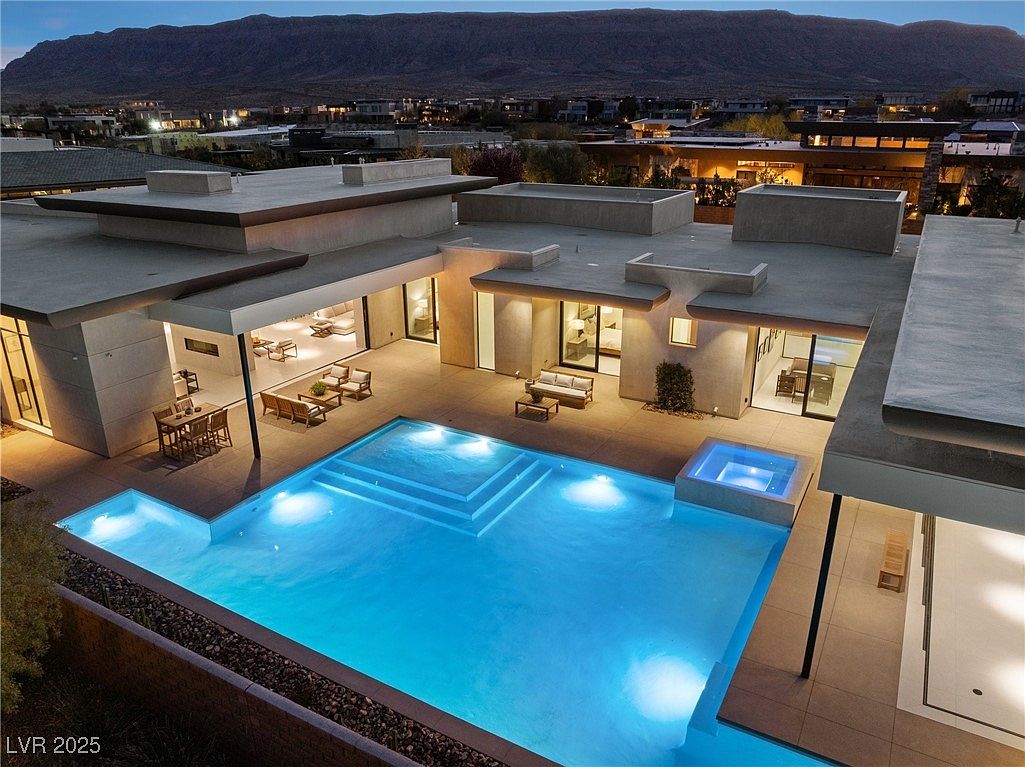 This is an aerial view of a modern luxury home featuring a large, illuminated swimming pool and spa. The pool has a multi-tiered entry and is surrounded by a spacious patio with outdoor seating areas. The architecture is contemporary with flat roofs and clean lines, set against a backdrop of distant mountains and city lights.