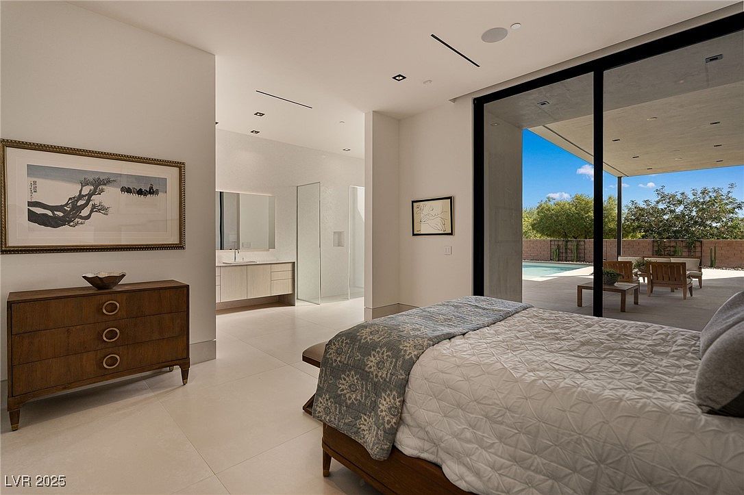 This is a view of a primary bedroom featuring a modern design with a seamless transition to an outdoor patio and pool area. The room includes a large bed with a patterned throw, a wooden dresser with artwork above, and an ensuite bathroom visible in the background. Large sliding glass doors provide ample natural light and a view of the outdoor living space.