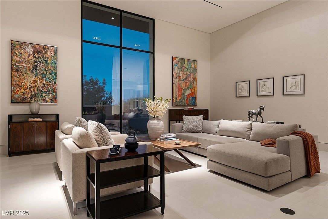 This is an interior shot of a modern living room, featuring a large sectional sofa, a side table with decorative items, and a coffee table with books and a vase of flowers. Artwork adorns the walls, and a large window provides natural light and a view of the outdoors. The room has a neutral color palette and a clean, contemporary design.