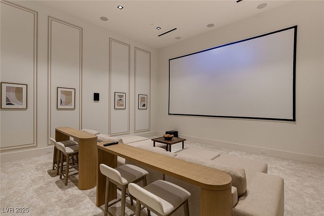 This interior shot showcases a modern home theater room, featuring a large projection screen, a low-profile sectional sofa, and a unique bar-height seating area with stools. The room is decorated in neutral tones with minimalist artwork, creating a sophisticated and comfortable entertainment space. The perspective is from the front of the room, capturing the full width of the screen and seating arrangement.