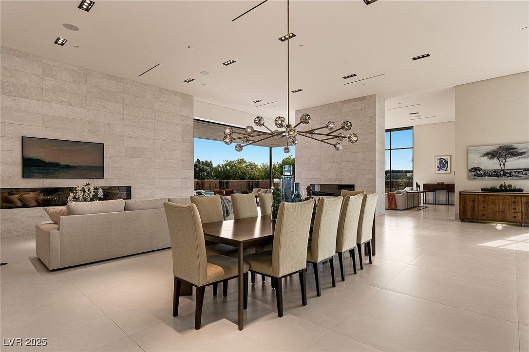 This is a spacious and modern dining room featuring a long wooden table with upholstered chairs, illuminated by a striking contemporary chandelier. The room boasts large windows offering natural light and views, complemented by a neutral color palette and sleek tile flooring. A fireplace and artwork add to the sophisticated ambiance, creating an inviting space for dining and entertaining.
