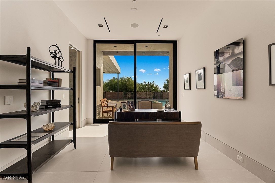 This is an interior shot of a modern office space. The room features a large sliding glass door that provides ample natural light and a view of the outdoor pool and patio area. The interior is decorated with contemporary art and a stylish bookshelf, creating a sophisticated and functional workspace.