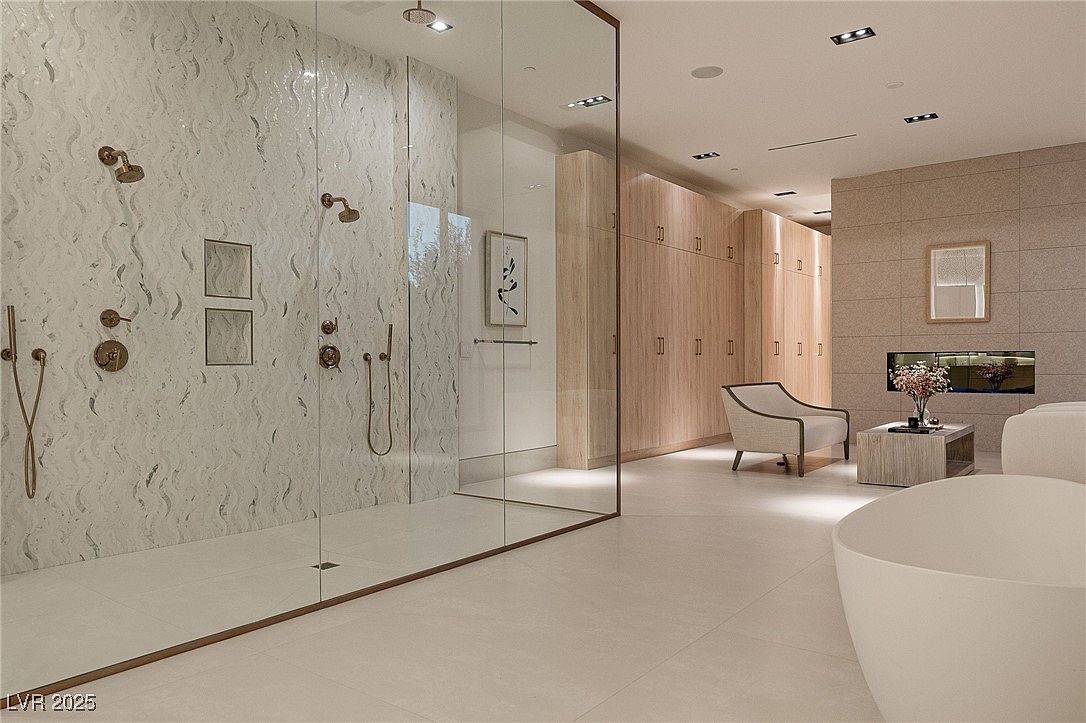 This is a luxurious primary bathroom featuring a spacious walk-in shower with dual shower heads and elegant tile work. The bathroom also includes a modern soaking tub, a seating area with a fireplace, and custom cabinetry for ample storage. The overall design is sophisticated and spa-like, creating a relaxing and upscale atmosphere.