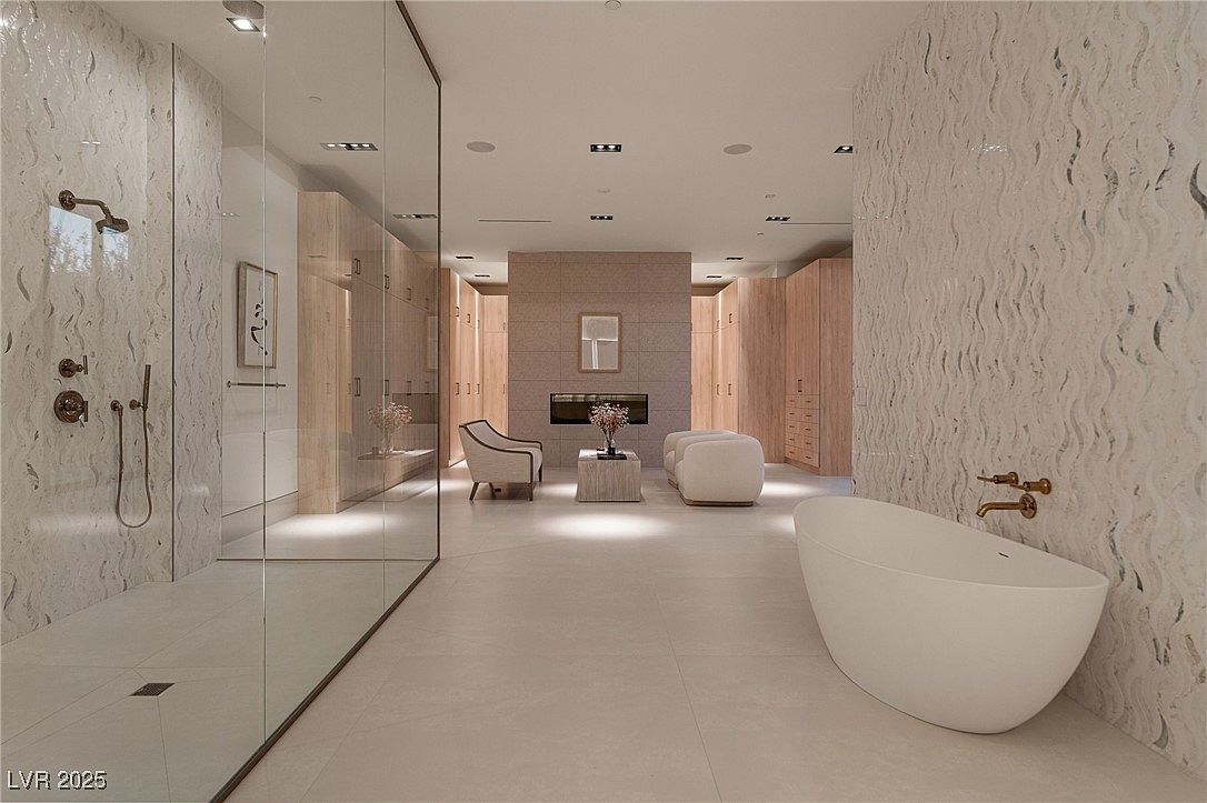 This is a luxurious primary bathroom featuring a glass-enclosed shower, a freestanding soaking tub, and custom wood cabinetry. The walls are adorned with textured tile, and the space includes a seating area with a fireplace, creating a spa-like atmosphere. The perspective is wide, showcasing the open layout and high-end finishes.