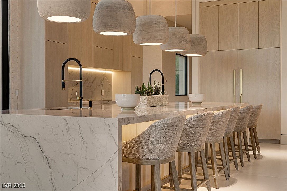 This is a modern kitchen featuring a large marble island with seating for six. The island has a black faucet and a decorative planter. The kitchen has light wood cabinets and pendant lighting above the island, creating a warm and inviting atmosphere.