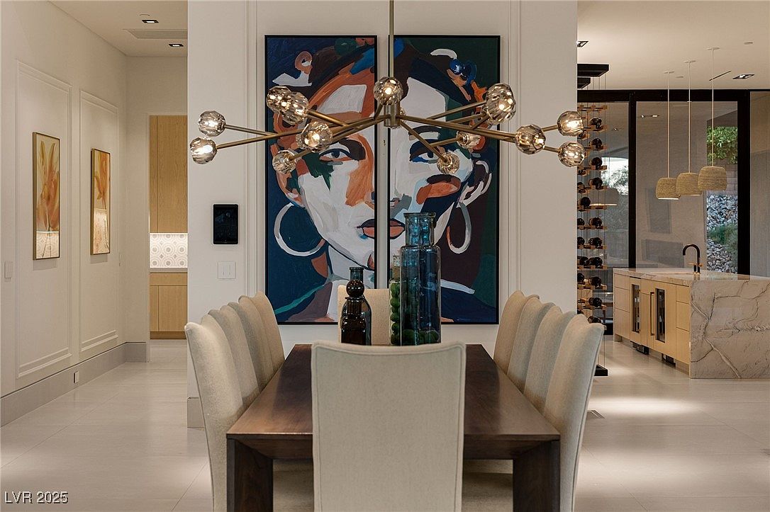 This interior shot showcases a modern dining room with a dark wood table surrounded by upholstered chairs. A striking chandelier hangs above the table, and a large diptych artwork featuring a portrait dominates the background. The room is bright and airy, with a glimpse into a wine cellar and kitchen area, suggesting a luxurious and open-concept living space.