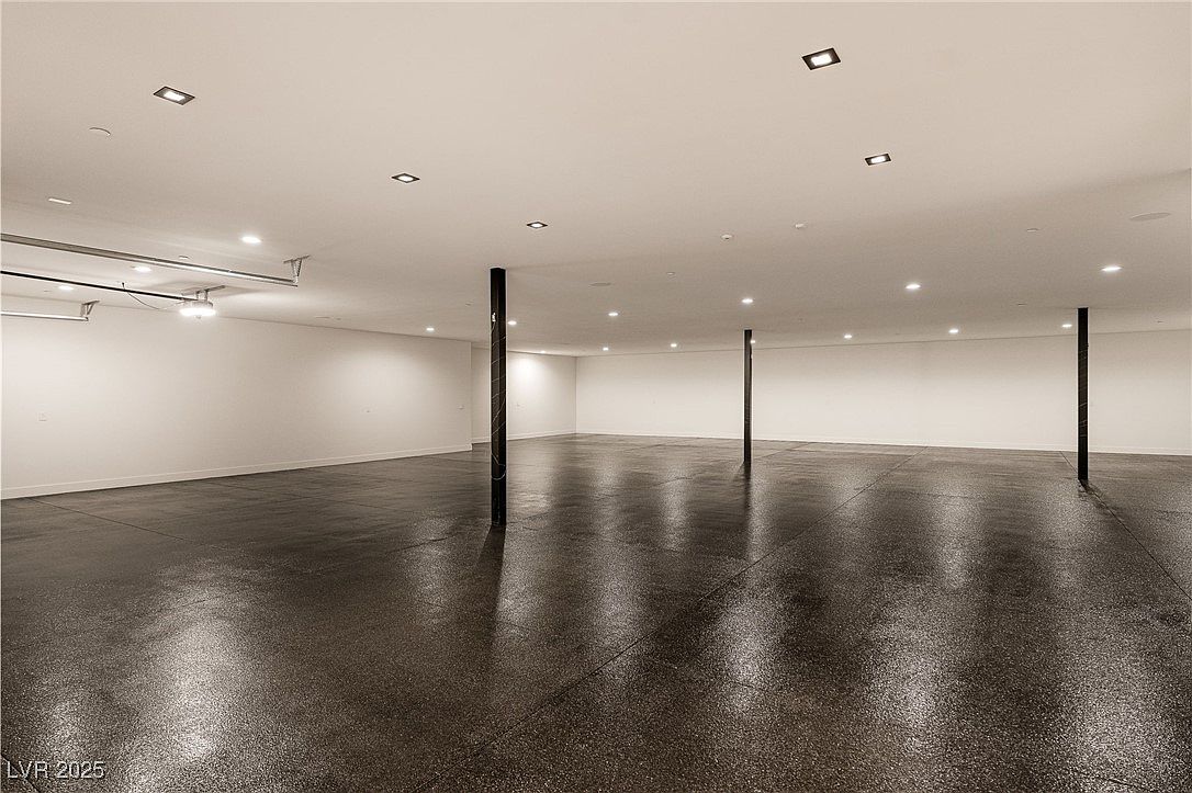 This is a spacious, unfinished basement with a polished concrete floor and white walls. The room is illuminated by recessed lighting and features support columns. The overall impression is clean and modern, offering a blank canvas for customization.