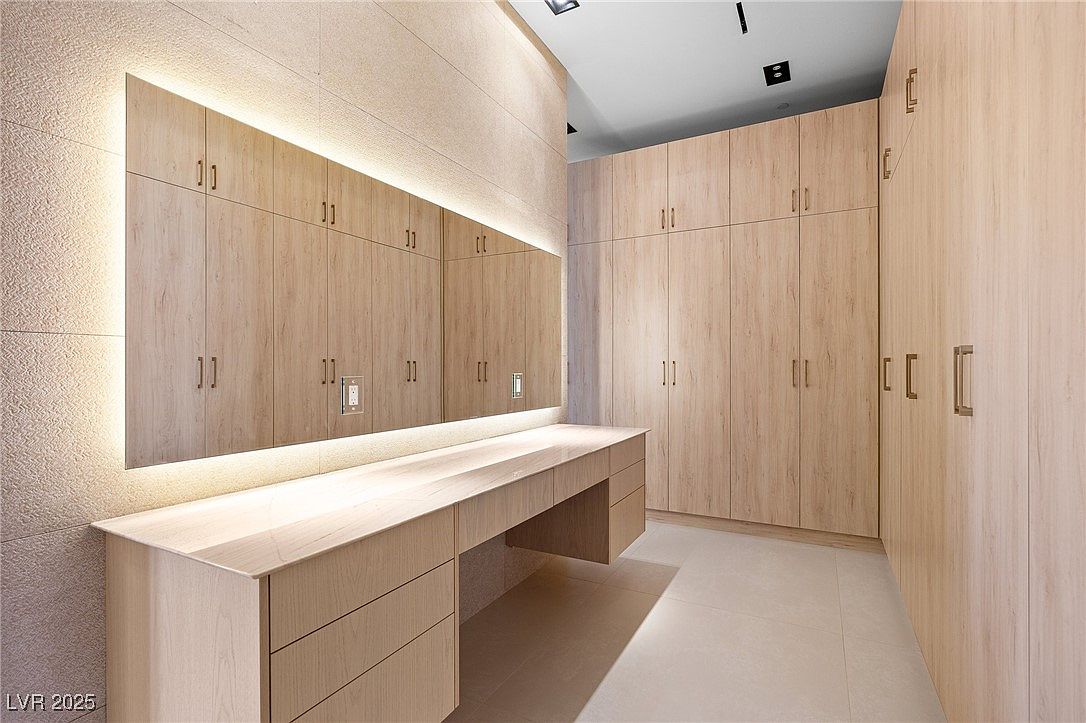 This is an interior shot of a well-organized walk-in closet. The closet features light wood cabinetry with sleek, minimalist hardware. A long vanity with a mirror and integrated lighting runs along one wall, providing ample space for dressing and grooming. The overall impression is one of luxury and functionality.