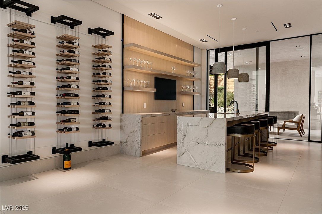This image showcases a modern wine cellar and bar area. The focal point is the wall-mounted wine racks displaying numerous bottles, complemented by a sleek bar counter with marble accents and stylish bar stools. The space is well-lit and features contemporary design elements, including floating shelves with glassware and a view to an outdoor area through large glass doors, creating an inviting and sophisticated atmosphere.
