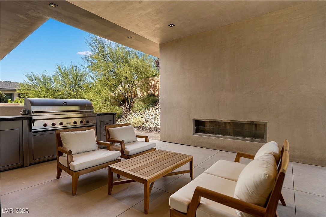 This image showcases a stylish outdoor patio area, featuring comfortable seating arrangements with wooden frames and light cushions, centered around a wooden coffee table. A built-in stainless steel grill and countertop area suggest an outdoor kitchen, while a modern fireplace adds a touch of elegance to the space. The patio offers a seamless transition between indoor and outdoor living, with views of the surrounding landscape.