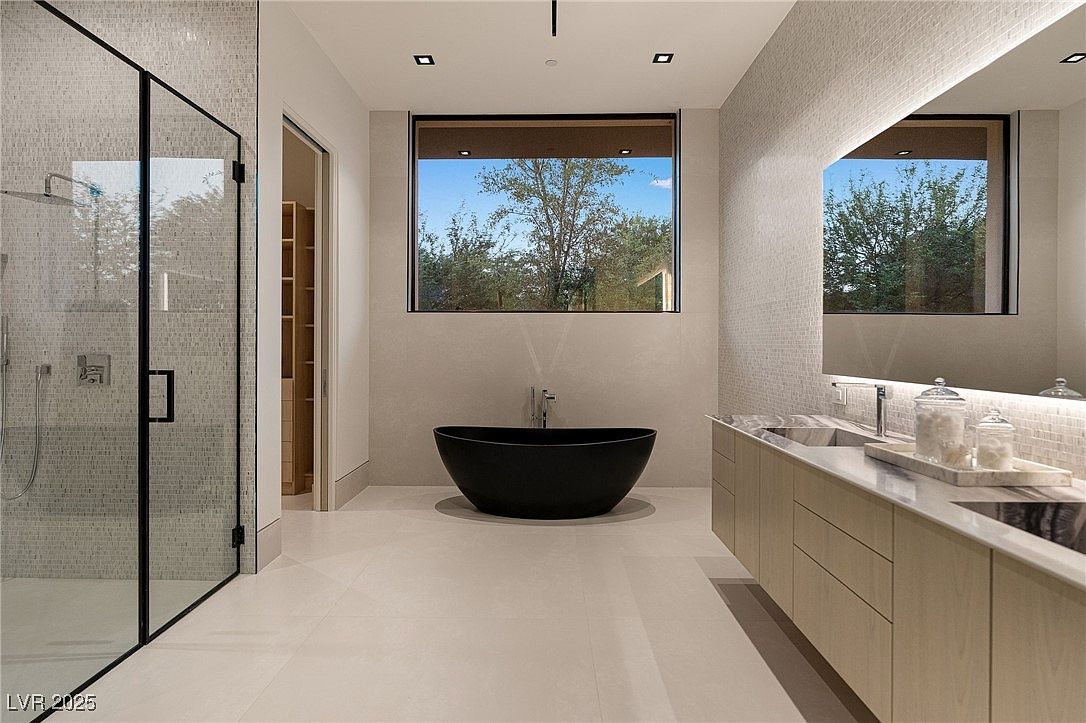 This is a modern primary bathroom featuring a glass-enclosed shower, a freestanding black bathtub under a large window, and a long vanity with light wood cabinets and a marble countertop. The walls are a mix of large format tiles and mosaic tiles, creating a luxurious and spa-like atmosphere. The perspective is from the doorway, showcasing the entire space.
