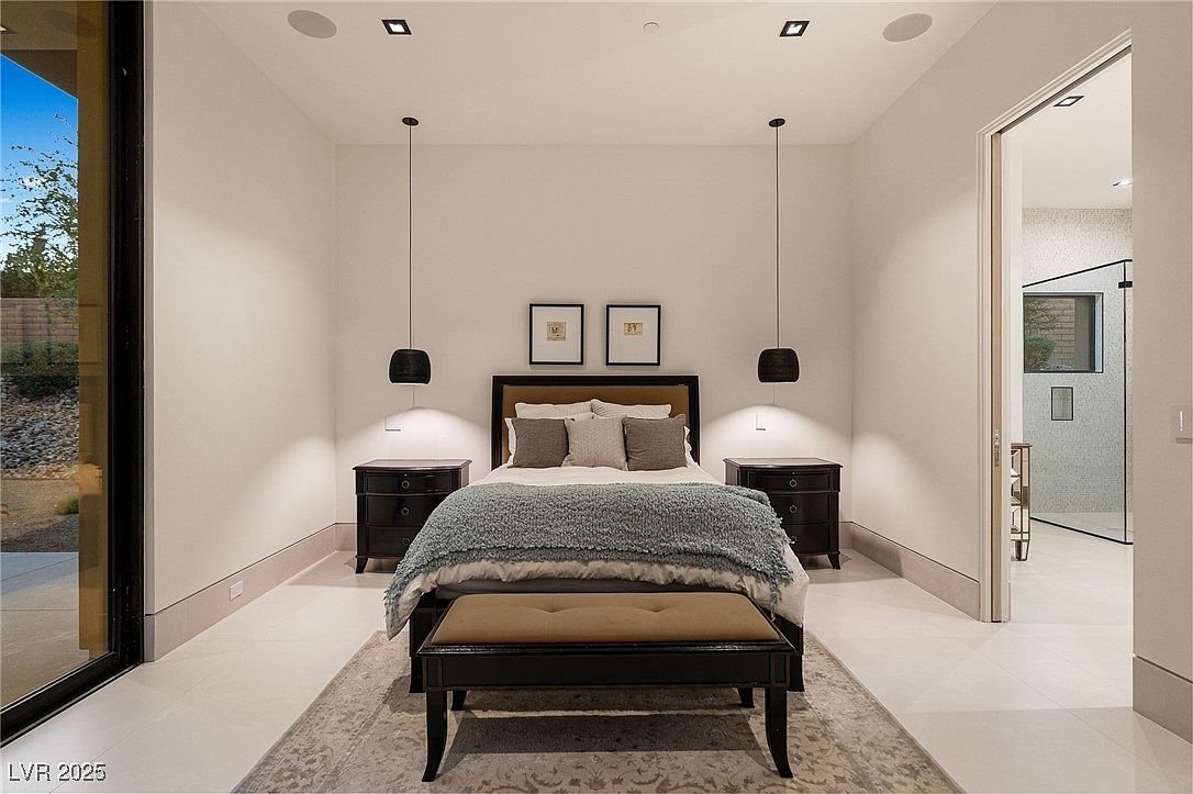 This is a well-lit primary bedroom featuring a bed with a padded headboard, flanked by matching dark wood nightstands and pendant lights. Two framed pictures hang above the bed, and a bench sits at the foot of the bed on top of an area rug. A doorway leads to an en-suite bathroom, enhancing the room's functionality and appeal.