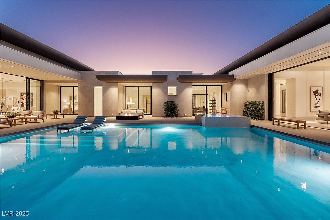 This image showcases a luxurious outdoor pool area at dusk, featuring a large, inviting pool with illuminated water. The modern architecture of the home surrounds the pool, with sliding glass doors leading to indoor living spaces. Lounge chairs are placed near the pool, creating a relaxing and upscale ambiance.
