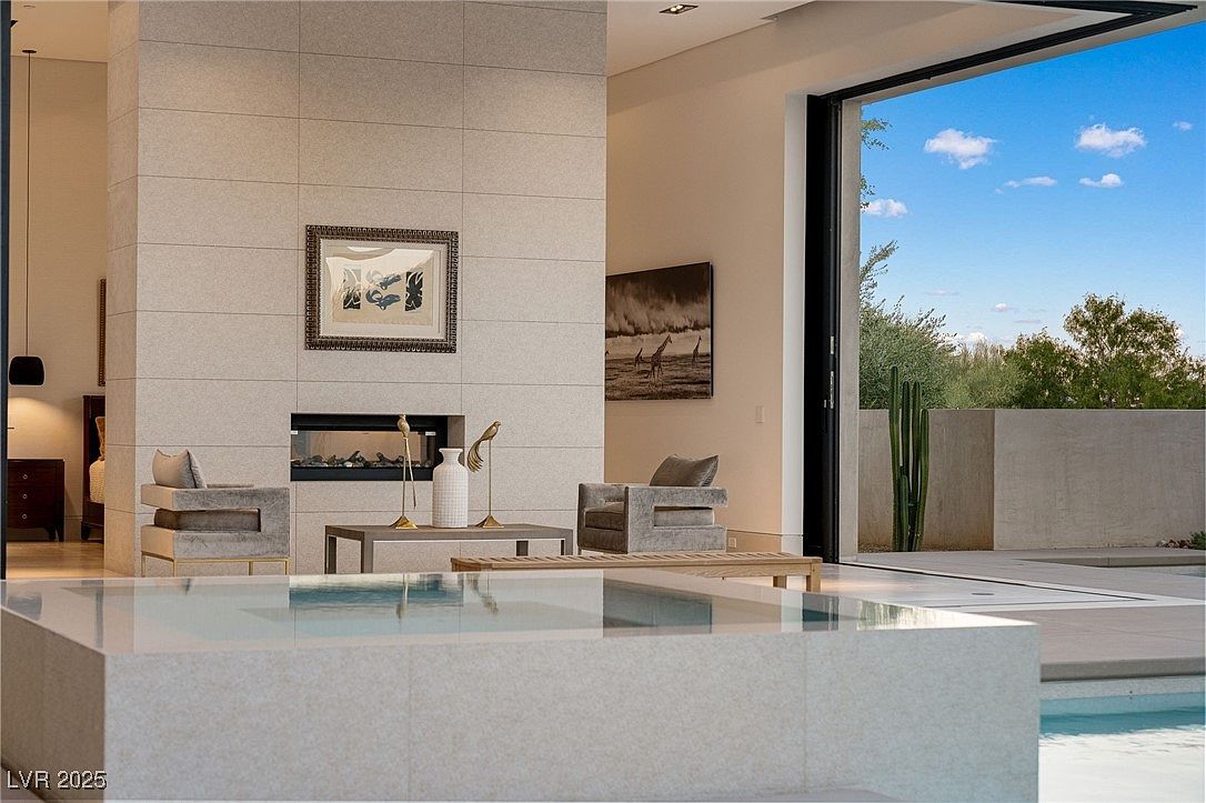 This interior shot showcases a modern living room with a focus on clean lines and neutral tones. A striking tiled fireplace wall anchors the space, complemented by contemporary seating and artwork. The room seamlessly transitions to an outdoor area through large sliding glass doors, offering a view of the pool and landscaping.