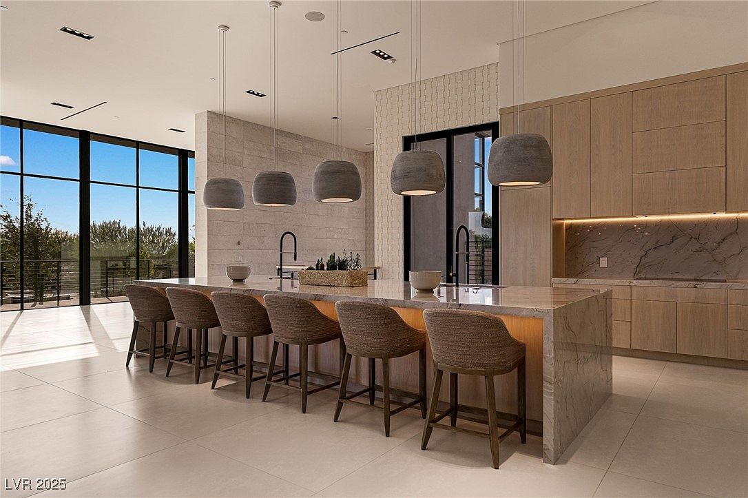 This is a modern kitchen with a large island featuring seating for at least eight people. The island has a marble countertop and is illuminated by pendant lights. The kitchen also features wooden cabinets and a large window with a view of the outdoors.