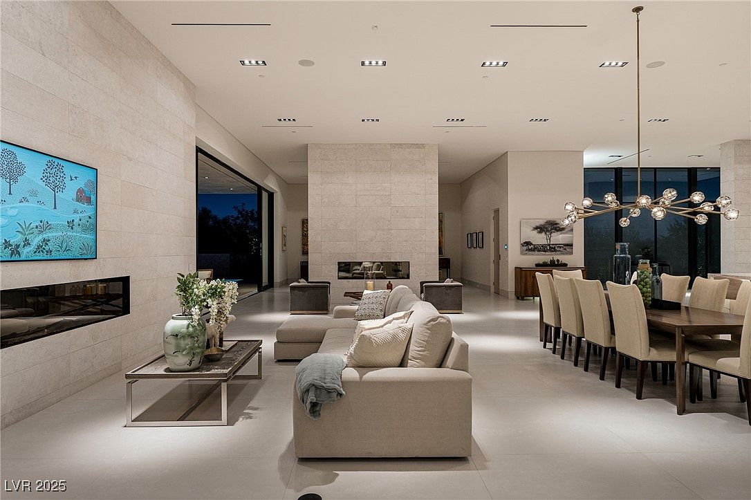 This is an interior shot of a modern living and dining area. The living room features a large sectional sofa, a coffee table with floral arrangements, and a fireplace with a television mounted above. The dining area includes a long wooden table with upholstered chairs and a modern chandelier, creating an open and luxurious space.