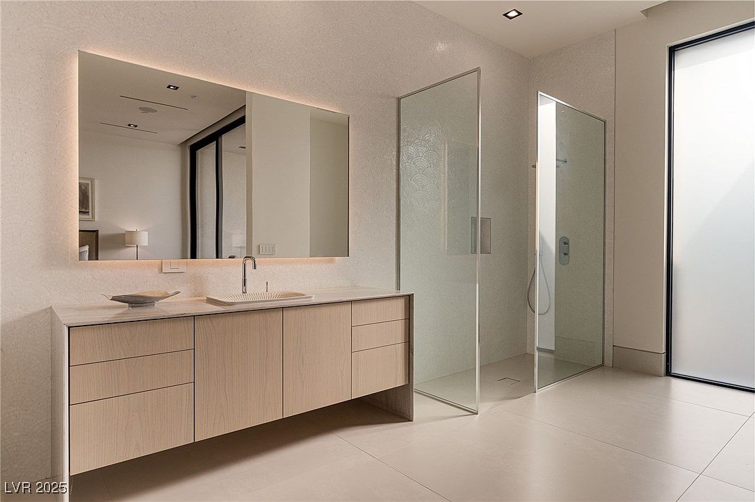 This is a modern primary bathroom featuring a floating vanity with light wood cabinetry and a large, illuminated mirror. The bathroom includes a glass-enclosed shower and a separate frosted glass door, contributing to a clean and minimalist aesthetic. The neutral color palette and tile flooring create a serene and luxurious atmosphere.