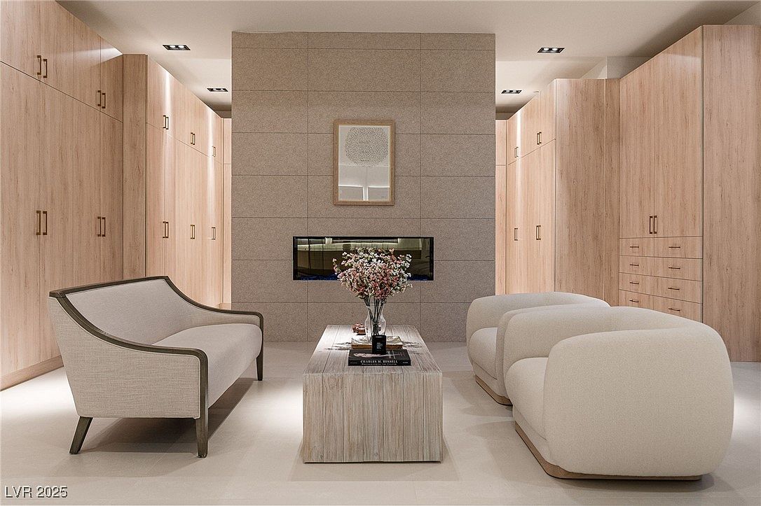 This interior shot showcases a modern living room with a neutral color palette. The room features a fireplace set within a tiled wall, flanked by built-in wooden cabinets. A light-colored sofa and two armchairs surround a wooden coffee table adorned with flowers and books, creating a serene and inviting atmosphere.