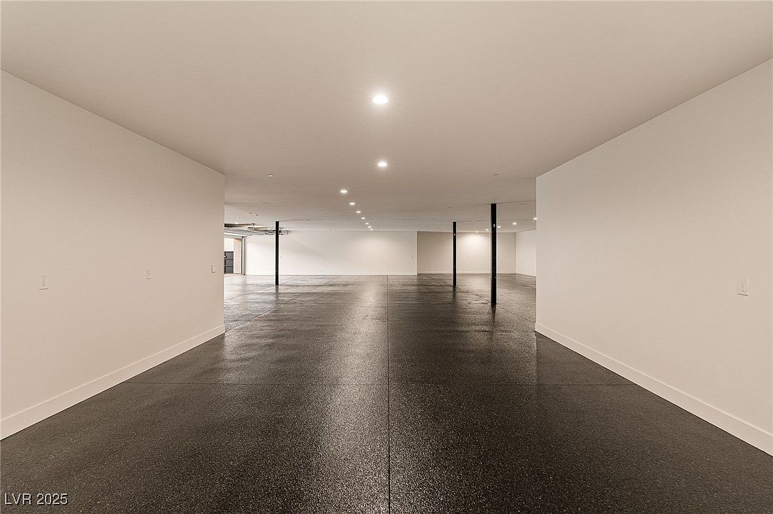 This is a spacious, unfinished basement with a dark, polished concrete floor and white walls. Black support beams are visible throughout the space, and recessed lighting illuminates the area. The room appears to be a blank canvas, ready for customization and finishing.