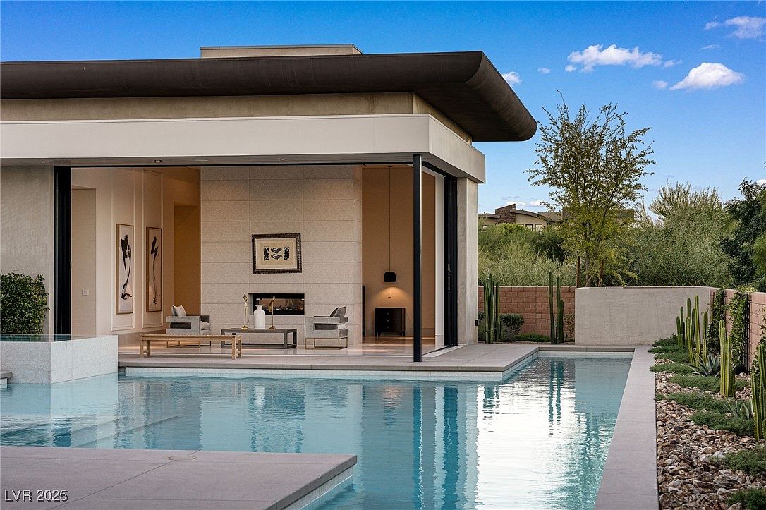This image showcases a luxurious outdoor pool area of a modern home. The pool's clear water reflects the contemporary architecture of the house, which features large sliding glass doors that open to an indoor living space. The landscaping includes minimalist plantings and decorative gravel, enhancing the sleek and sophisticated aesthetic.