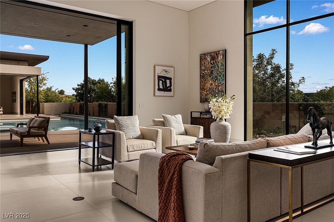 This is an interior shot of a modern living room featuring neutral tones and clean lines. The room is well-lit with natural light streaming through large windows, offering views of the outdoor pool and greenery. The furniture includes a sectional sofa, armchairs, and a console table, creating a comfortable and stylish living space.
