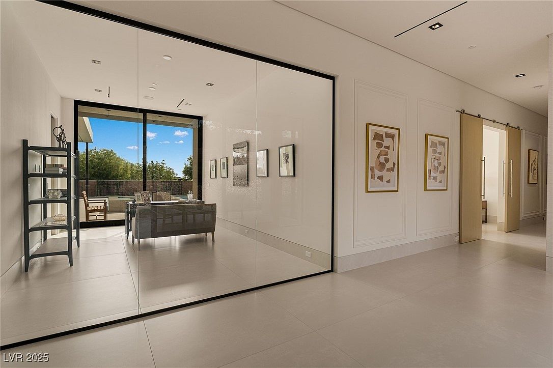 This interior shot showcases a modern hallway with large format tile flooring and minimalist design. A large glass wall reflects a living area, creating an illusion of expanded space. The hallway features framed artwork and a sliding barn door, adding a touch of contemporary elegance.