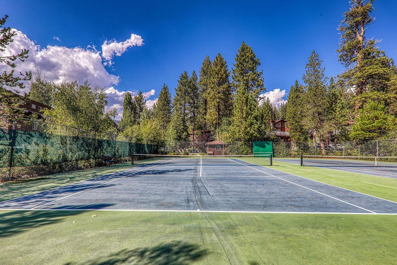 This image captures a well-maintained outdoor tennis court facility surrounded by a chain-link fence and lush pine trees. The court features a blue playing surface with white lines, set against a backdrop of a clear blue sky with scattered clouds. The perspective is centered from the baseline, providing a clear view of the net and the surrounding recreational environment.