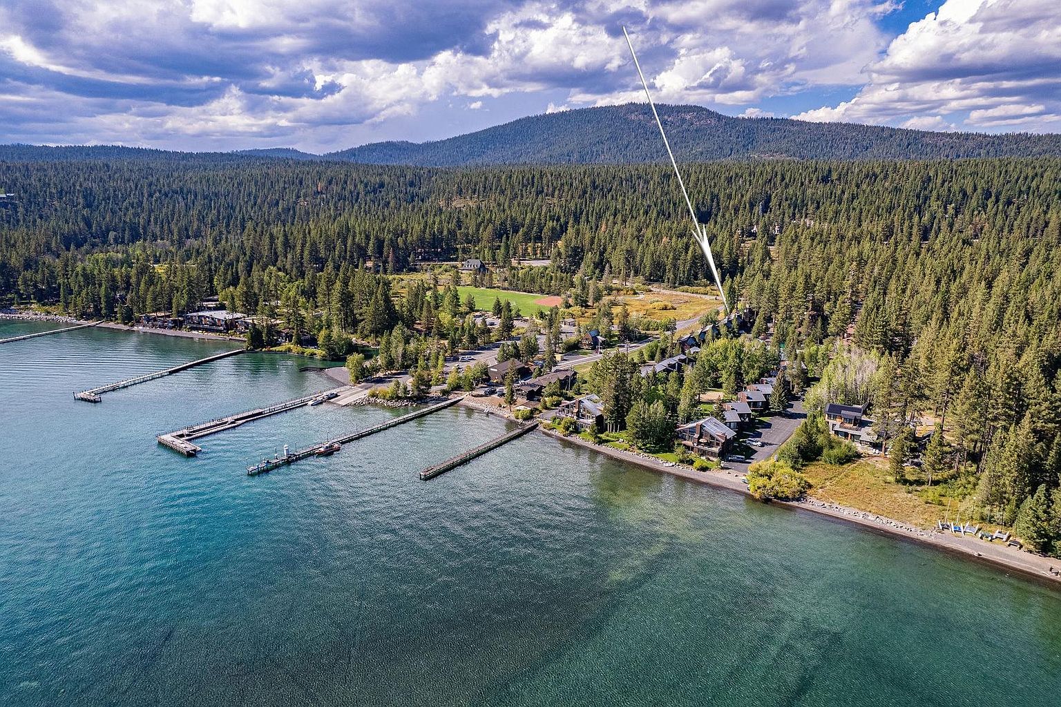 This high-angle aerial perspective captures a scenic lakeside community nestled against a dense, forested mountain backdrop. The image highlights the property's proximity to the water, featuring multiple wooden piers extending into the clear lake and a cluster of residential buildings surrounded by mature trees. A white arrow points toward a specific unit within the complex, emphasizing its location within this tranquil, nature-focused setting.