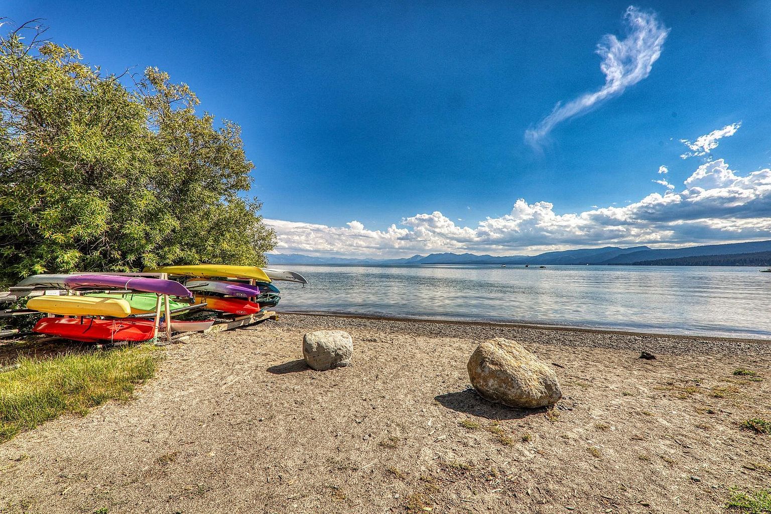 This scenic waterfront area features a sandy beach shoreline with a rack of colorful kayaks stored near a lush, green tree. Two large, weathered boulders sit prominently on the sand, leading the eye toward the expansive lake and distant mountain range under a bright blue sky with scattered clouds. The perspective is a wide-angle, eye-level shot that captures the serene and recreational atmosphere of the property's lakefront access.