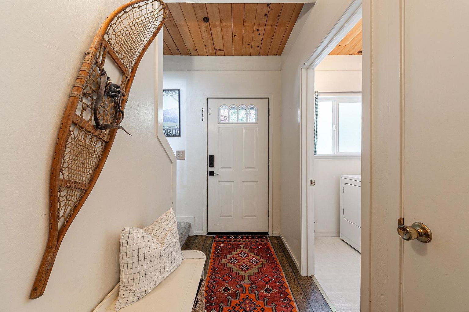 This inviting entryway features a white front door with a decorative glass insert, complemented by a warm wood-paneled ceiling. A vintage-style snowshoe is mounted on the wall above a white bench with a patterned pillow, while a vibrant, colorful runner rug adds character to the dark wood flooring. To the right, an open doorway provides a glimpse into a laundry area, creating a functional and welcoming transition space.